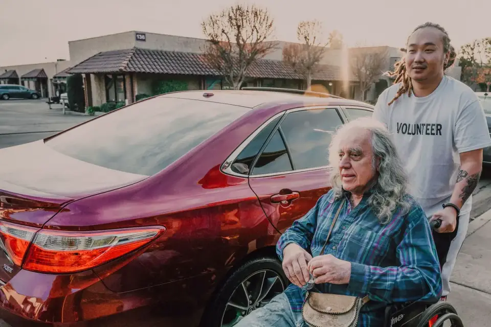 A driver holding a car door open while a senior dog is helped into the back seat
